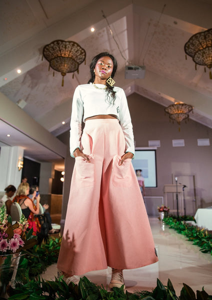 Model Stands at the end of a runway her dark skin shining in the spotlight as the crowd applauds. She is wearing one large gold GNK earring, with a white Chilli Crop Skivvy in organic cotton and the Jen Pant flares in Pink cotton. She faces the camera with a smile, her hands resting in the front pockets of the pants.
