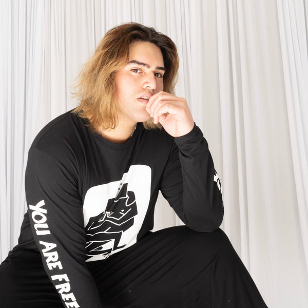 Sam wears a black long sleeve tee shirt with the Two Spirits print in white ink depicting two angular human figures facing each other with their arms and legs intertwined with one another. On the left sleeve is reads 'Genkstasy' down the arm, on the right sleeve it reads 'You are free' with the Genkstasy logo at the wrist. Sam is looking at the camera with a dreamy look of contemplation.
