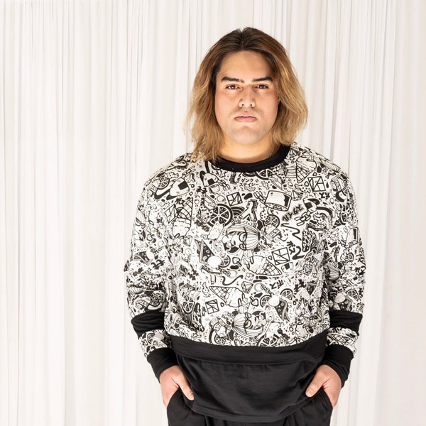 Sam wears the GNKY Jumper and GNKY Track Pants, he's looking straight at the camera and is standing face on with his hands in his pants pockets. The jumper is printed in black and white graphic style, with black cuffs at the wrist and bands half way between the wrist and elbow, as well as a black collar trim and hem band. The jumper has long sleeves and is a cropped length sitting just above high hip on a tall human like Same who is 195cm tall, or at the hips on a shorter human.