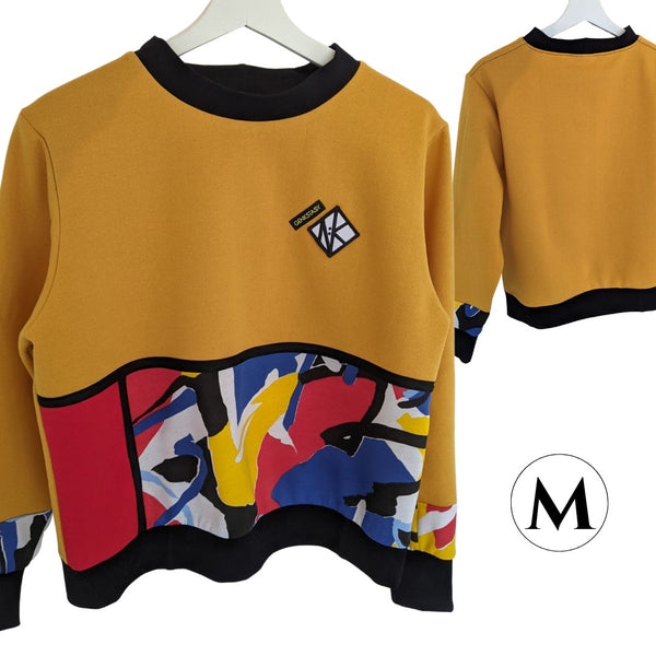 Size M, which is mainly rich gold ochre, with Mondrian panel details bordered in black around a red panel and a patterned panel from the waist to the hem cuff. It is a fleecey lined jumper, cropped at the hip for ease of wearing in a wheelchair, or showing off your booty. The cuffs are well fitted and 8cm long, and they and the collar and hem of the jumper are black cotton for comfort and contrast with the geometric colourful pattern of the bottom panel.