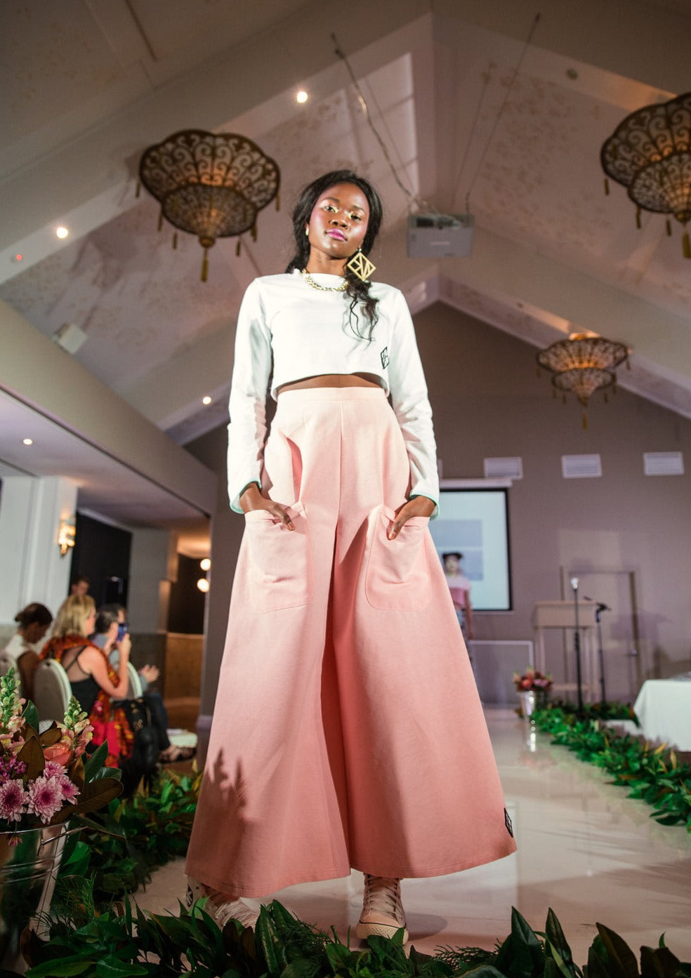 Model Stands at the end of a runway her dark skin shining in the spotlight as the crowd applauds. She is wearing one large gold GNK earring, with a white Chilli Crop Skivvy in organic cotton and the Jen Pant flares in Pink cotton. She faces the camera with a smile, her hands resting in the front pockets of the pants.
