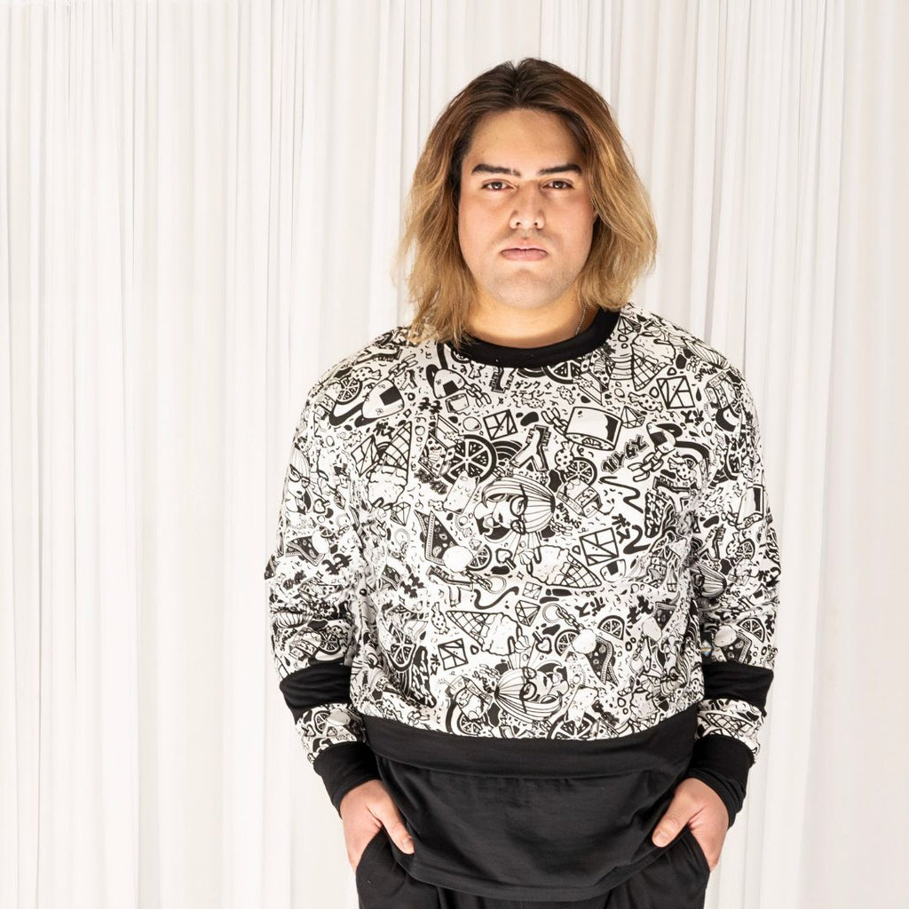 Sam wears the GNKY Jumper and GNKY Track Pants, he's looking straight at the camera and is standing face on with his hands in his pants pockets. The jumper is printed in black and white graphic style, with black cuffs at the wrist and bands half way between the wrist and elbow, as well as a black collar trim and hem band. The jumper has long sleeves and is a cropped length sitting just above high hip on a tall human like Same who is 195cm tall, or at the hips on a shorter human.