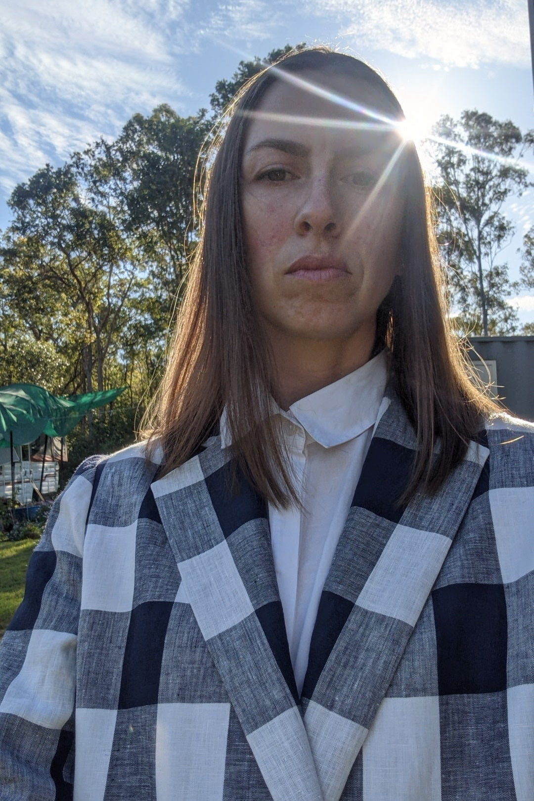 Person wearing a checked linen blazer with white crisp shirt underneath. The image is a close up showing the detail of the wide lapels, and there are trees and sunlight in the background