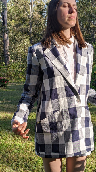 Model standing outdoors in a well made checkered linen jacket, a crisp white collared shirt underneath in front of a verdant natural backdrop. The Jacket has large angle-cut pockets, and two buttons in the front to fasten it.