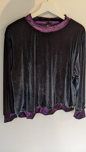 This is the front view of the Velvet Tidal Jumper in size XL, hanging on a hanger. The Velvet is almost black with a turquoise sheen, with Amythyst purple hem, collar, and sleeve cuffs. On the hem at the right hand side is a small Genkstasy logo tag on the outside of the purple hem. The tag is black with gold embroidery to show the geometric Genkstasy logo.