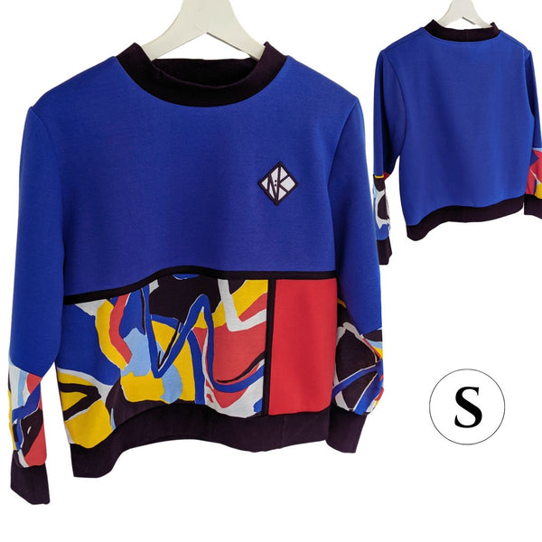 The Tidal Jumper in size Small, with Mondrian panel details with black borders around a red panel and a patterned panel from the waist to the hem cuff. It is a fleecey lined jumper in royal blue, and is a slightly boxy fit, cropped at the hip for ease of wearing in a wheelchair, or showing off your booty. The cuffs are well fitted and 8cm long, and they and the collar and hem of the jumper are black cotton for comfort and contrast with the geometric colourful pattern of the bottom panel. 