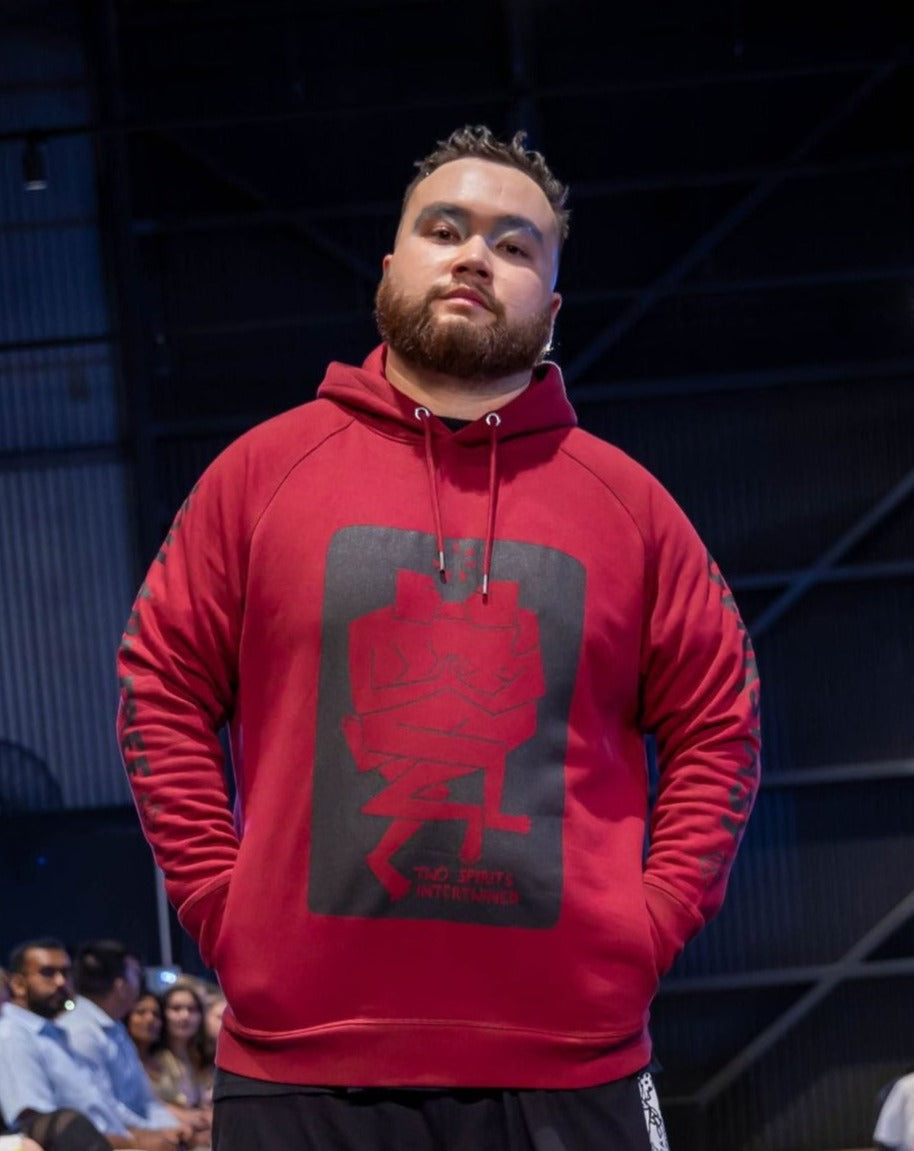 Man stands proudly with his hands in the pockets of his rich burgandy red hooded jumper. On the front of the jumper is a black graphic art print of two figures intertwined together. It reads Two Spirits Intertwined. On the sleeves it reads 'You Are Free' on one, and 'Genkskstasy' on the other.