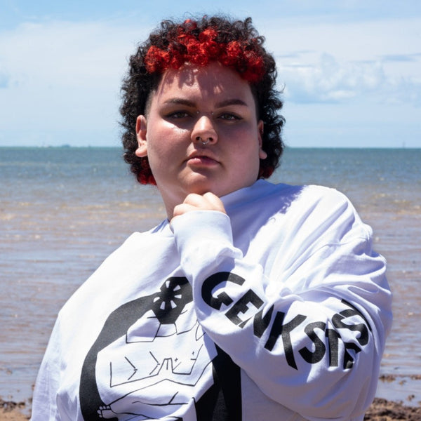 Ash is wearing the Two Spirits organic cotton tee in white with black print. They have facial piercings and defined brows, and they're holding the soft shirt collar up to their neck so you can see the word Genkstasy (joyful spirit) printed on the sleeve. They are at the beach and their red and black curls are shining in the sun as they look straight at us with both strength and softness.